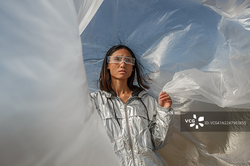 身着银色未来感服装的年轻亚洲女子，在风中被半透明塑料环绕图片素材