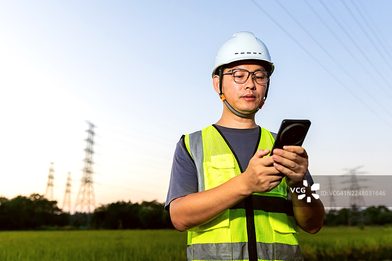 一名亚洲工程师在高压电塔下使用手机。图片素材