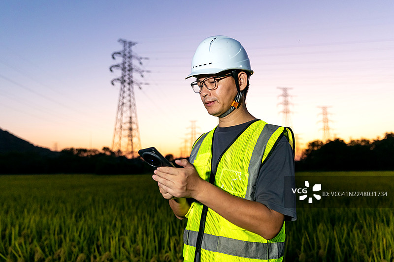 一位亚洲工程师在高压电塔下使用手机。图片素材