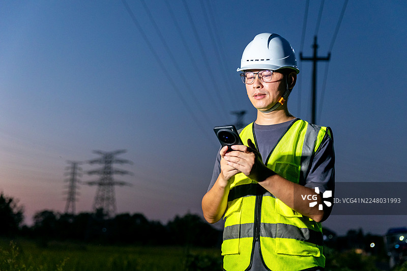 一位亚洲工程师在高压电塔下使用手机。图片素材