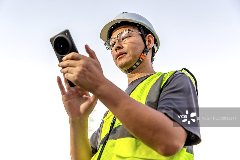 一位亚洲工程师在高压电塔下使用手机。图片素材