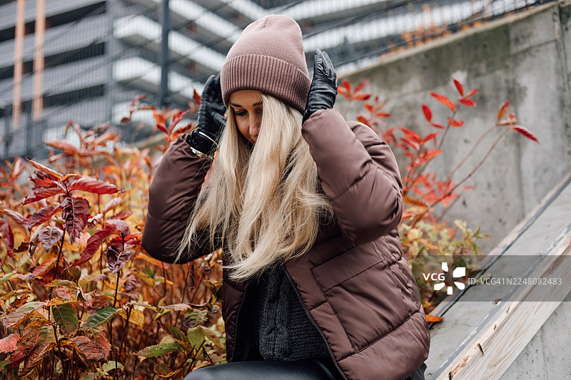秋日街景：戴着棕色羽绒服的女士在红叶间调整帽子图片素材