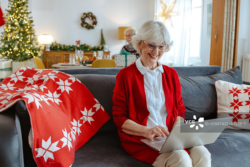 一位年长女性在装饰圣诞节的客厅沙发上使用笔记本电脑，享受欢乐的节日时光图片素材