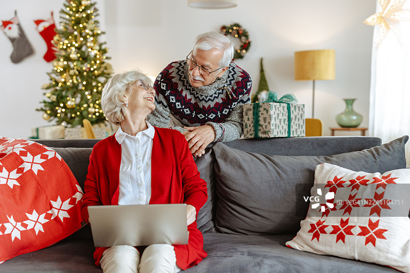 一对老年夫妇在家中享受圣诞节，有笔记本电脑、礼物和节日装饰。图片素材