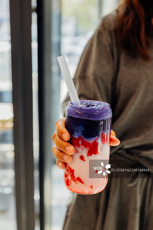 白色桌上，女性的手拿着一杯冰镇蝴蝶抹茶拿铁，旁边还有覆盆子冰沙。一杯冰爽的夏日饮品。无咖啡因饮品。健康食品概念。图片素材