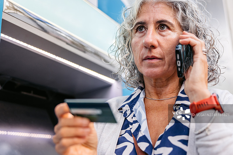 一位成熟的女士在西班牙巴塞罗那的自动取款机前，一边拿着信用卡一边打电话。图片素材