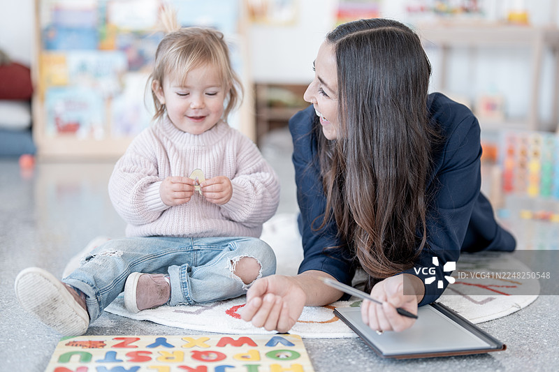 看护者与小女孩在家中学习，平板电脑和字母拼图在地板上图片素材