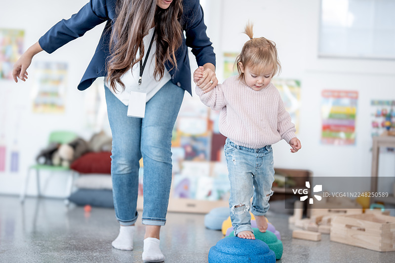 慈爱的女性在明亮的玩耍室里，一边玩耍，一边帮助蹒跚学步的幼儿保持平衡并进行探索图片素材