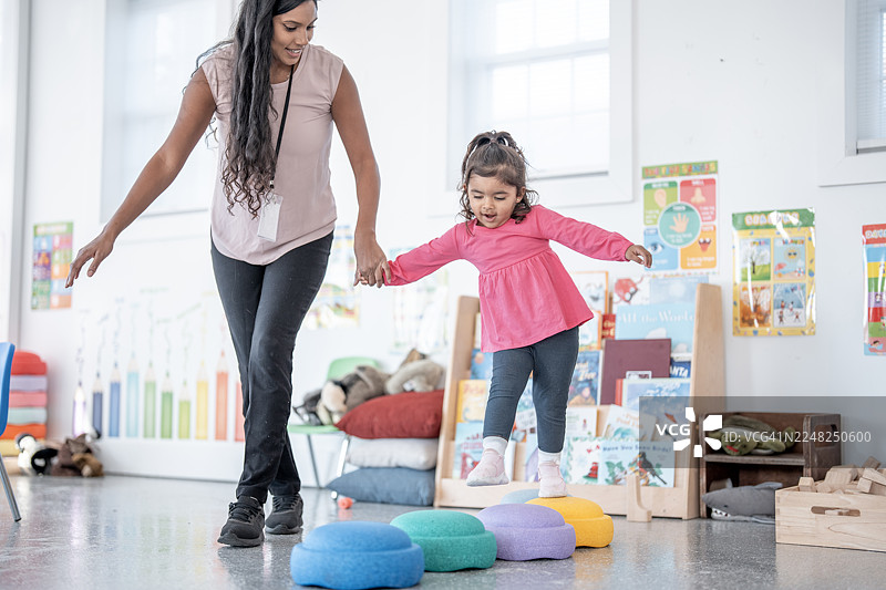 一位有色人种女老师指导一个小女孩跨越彩色踏脚石，教室里充满欢乐的瞬间图片素材