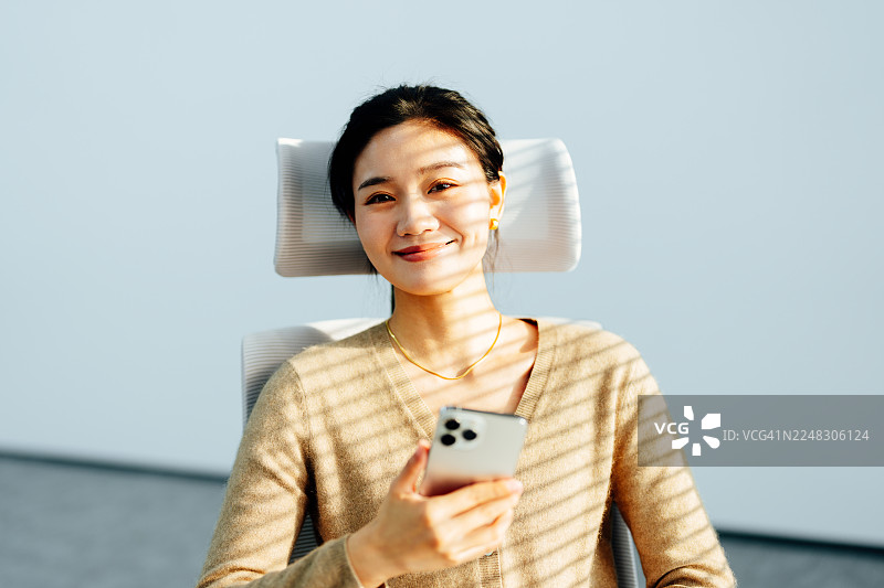 年轻的亚洲女商人坐在办公室，在窗帘阴影下，手持智能手机，看向镜头。在线学习。商业规划。橙色石头。图片素材