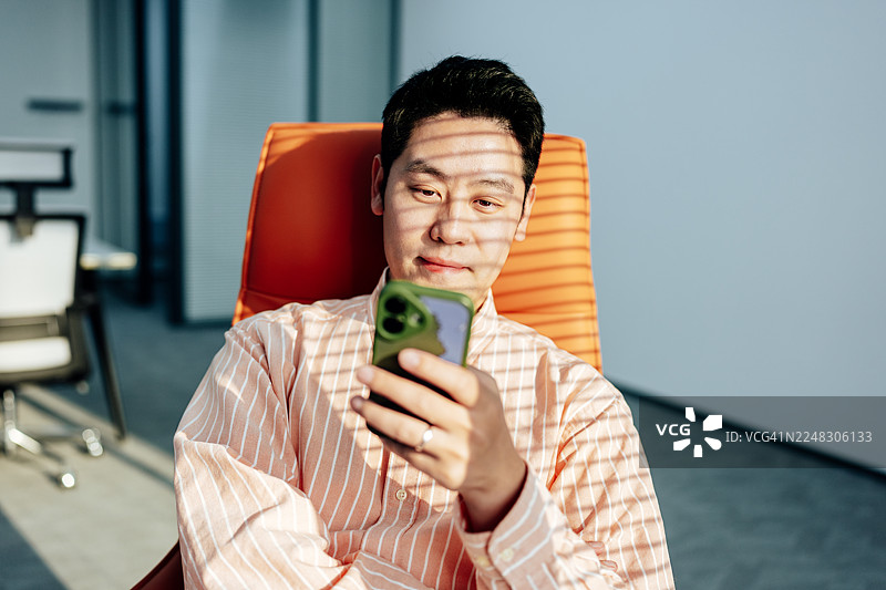 年轻的亚洲商人坐在办公室的橙色沙发上，窗帘的阴影投射下来，他正看着智能手机。橙色调。在线学习。商业规划。图片素材