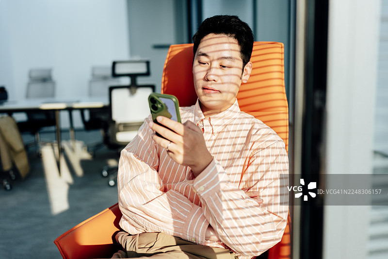 年轻的亚洲商人坐在办公室橙色沙发上，窗帘投下的阴影下，他正在使用智能手机。橙色调。在线学习。商业规划。图片素材