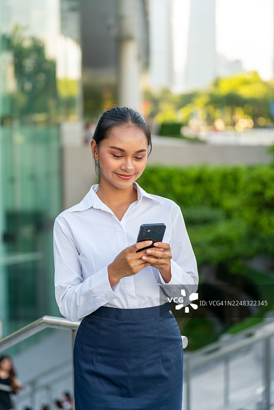 亚洲女商人的肖像，她在现代城市的户外使用手机。一名微笑的亚洲女商人正在城市区域的户外用手机发短信。图片素材
