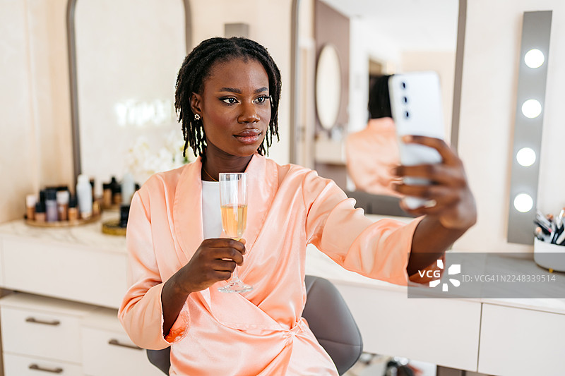 年轻的黑人女性在美容院化好妆后，一边喝香槟一边用手机自拍图片素材