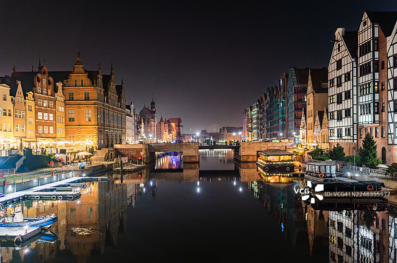 夜幕下的格但斯克老城河畔，历史建筑灯火辉煌，平静的水面映照出清晰的倒影，泊岸的船只和延伸的滨水区温暖的街灯交相辉映。图片素材