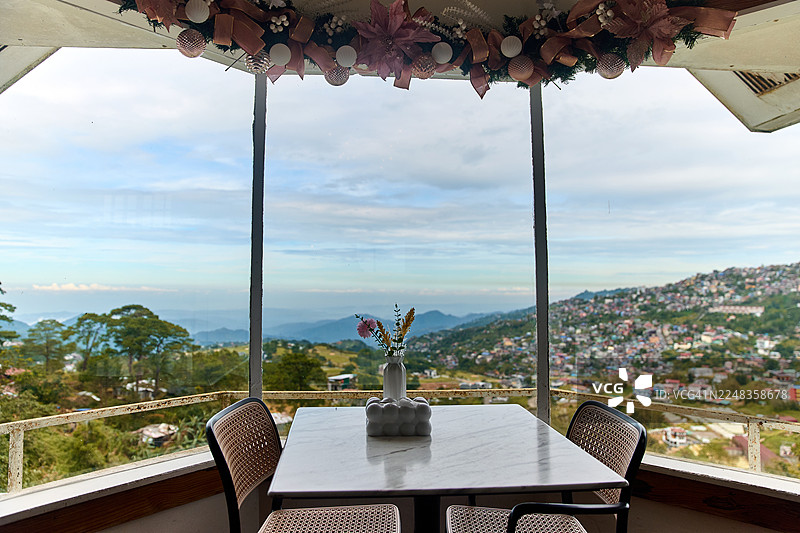 窗边摆放着一张桌子、两把椅子和一个花瓶，窗外是色彩斑斓的山坡城镇和远处的群山。图片素材