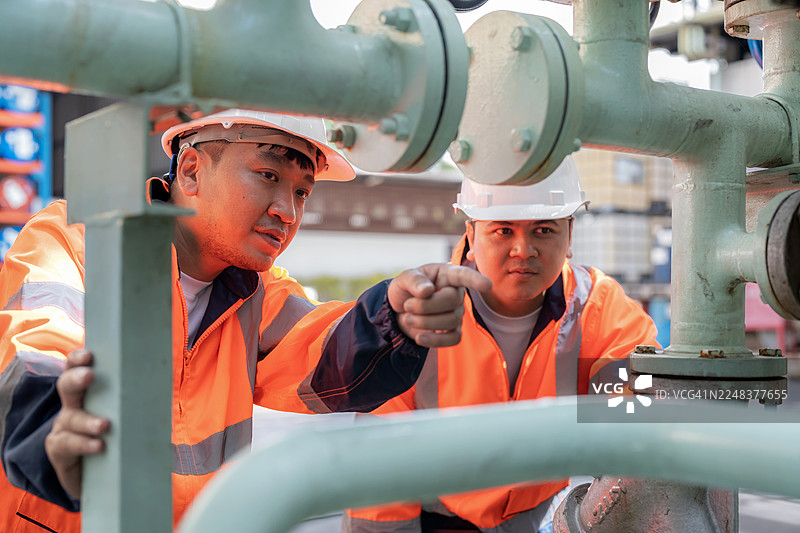 两位全副武装的安全工程师正在润滑油生产线上检查和调整一个输油管道系统。他们正在石化工厂讨论机器阀门设备润滑工作流程和系统性能，涉及高科技。图片素材