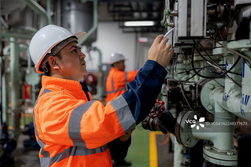 一位身着防护装备的机械工程师在炼油厂中，一边监控仪表确保阀门处于正常范围内，一边调整压力系统参数，并使用数字平板同步数据，以提高效率和安全。图片素材