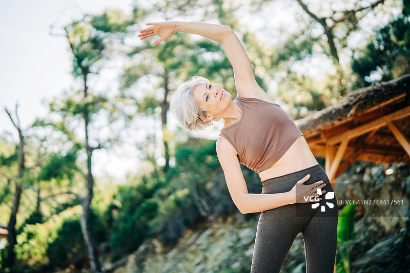 一位健美的女性在宁静的湖边伸展手臂，背景是绿色的山丘。这个户外锻炼场景体现了健康、平衡以及在大自然中积极健康的生活方式。图片素材