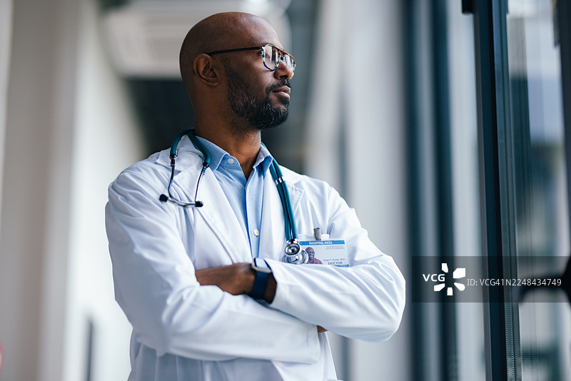 一位身穿白大褂、佩戴听诊器的医生，自信地站在医院走廊，眺望窗外。图片素材