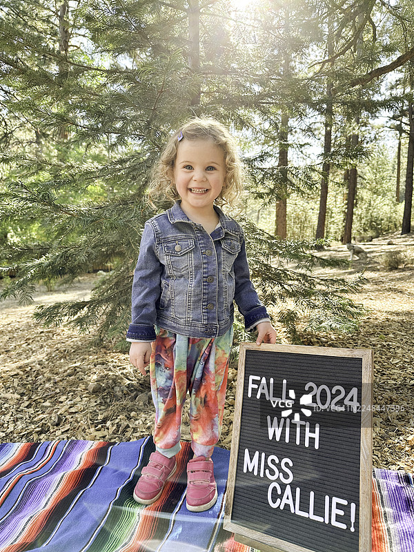 一个可爱的学龄前儿童站在户外，背景是美丽的风景，面前是“开学第一天”的标志。图片素材