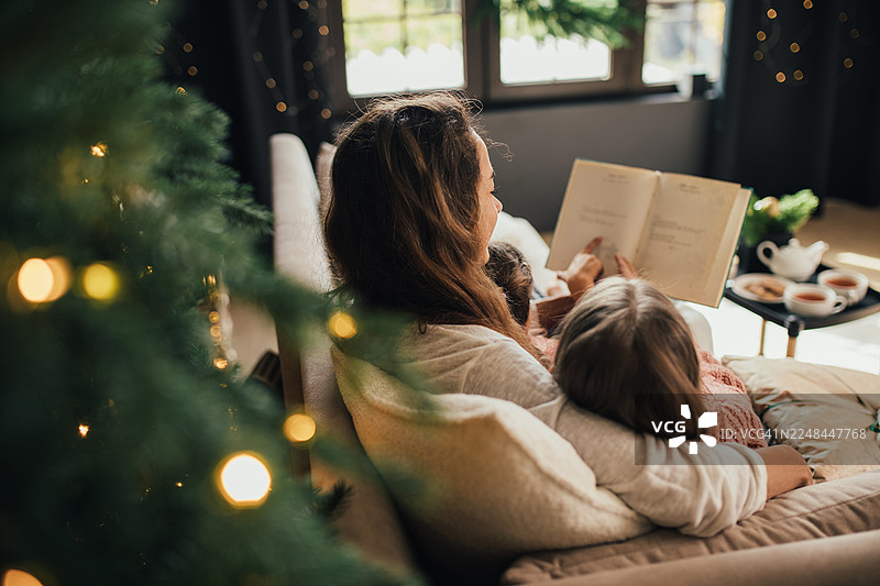 温馨的起居室内，妈妈和孩子在圣诞树旁舒适地依偎在沙发上阅读图片素材