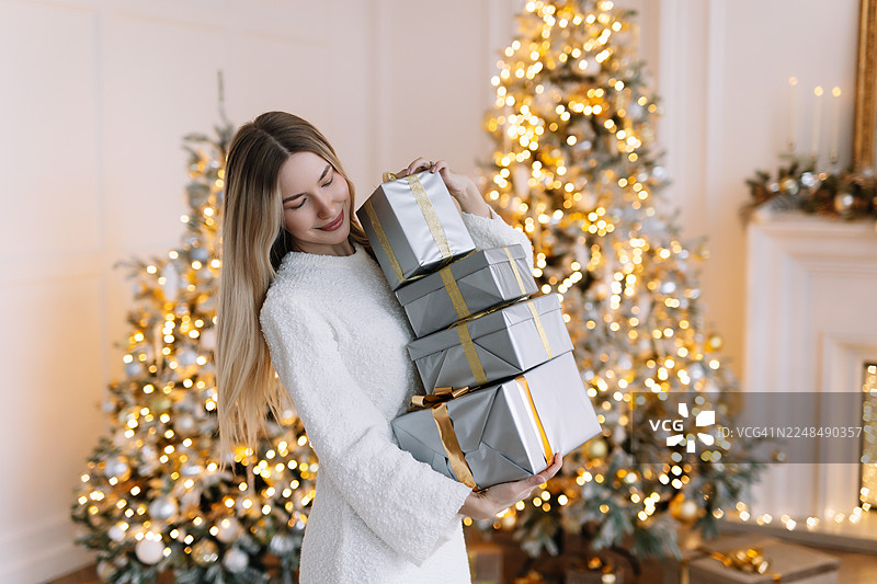 身穿白色长裙的年轻女子，脸上带着快乐的笑容，手里拿着一堆圣诞礼物，坐在舒适客厅的沙发上，旁边是装饰好的新年季节的圣诞树，在家中享受节日时光。图片素材