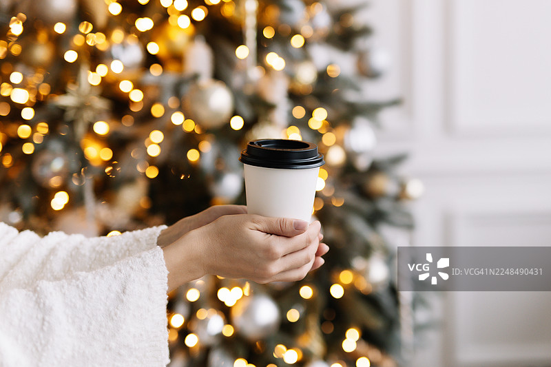 十二月新年假日冬季，在装饰好的圣诞树和闪烁的灯饰旁，一双人类的手正捧着一杯热咖啡，体现了圣诞饮品的概念。图片素材