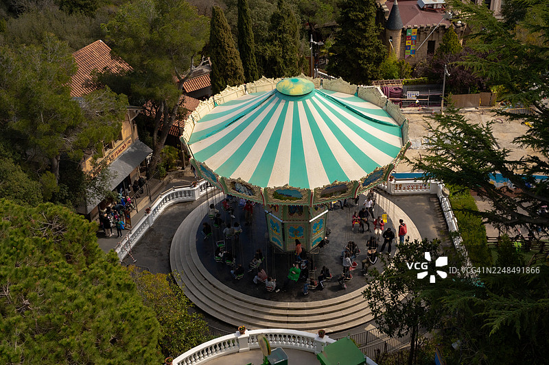 俯视角度拍摄的巴塞罗那蒂比达博游乐园的彩色旋转木马，上面有游客，周围环绕着松树图片素材