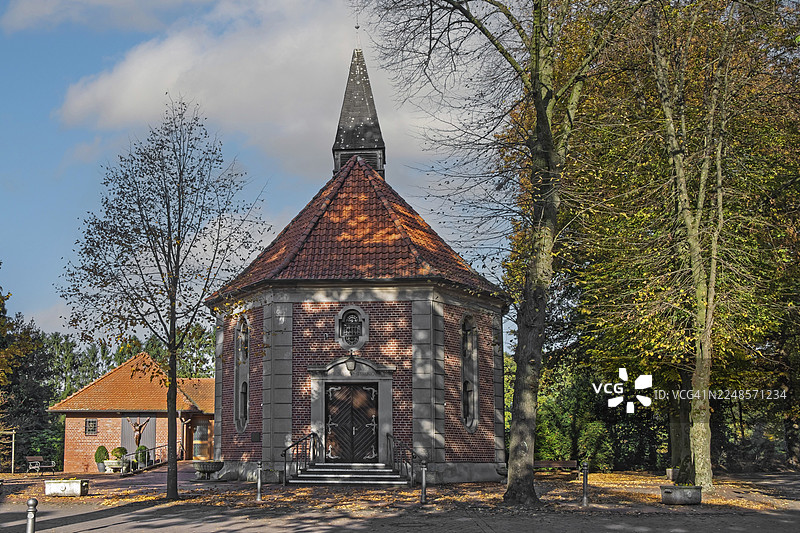 一座砖砌教堂，有尖顶和屋顶尖塔，周围是秋天的树木，背景是蓝天，位于德国北莱茵-威斯特法伦州明斯特兰的希克-阿勒。图片素材
