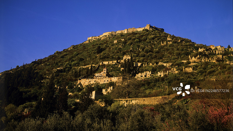 坐落于长满杂草的山丘上的废墟，背景是深邃的蓝色傍晚天空，位于希腊伯罗奔尼撒半岛的斯巴达西北部、塔耶托斯山脉山麓的米斯特拉斯（拜占庭中世纪遗址，联合国教科文组织世界遗产地）。图片素材