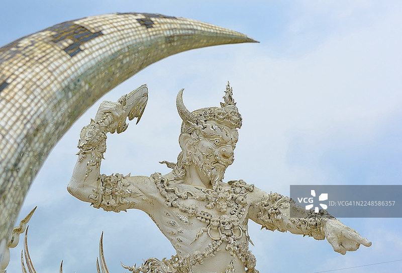 泰国蓝天下，一座白色神话生物雕像，头上有角，一只手臂举向马赛克拱门图片素材