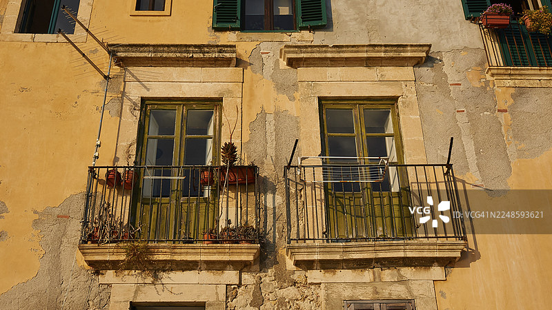 阳光下，有两扇阳台、窗户和鲜花的饱经风霜的历史建筑外墙，位于意大利西西里岛锡拉库扎的奥提伽岛历史中心。图片素材