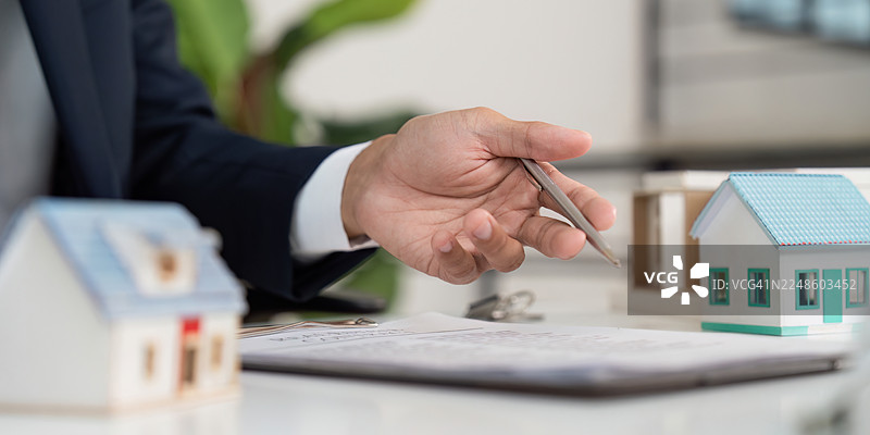 一位商人正在办公桌前，手中拿着笔，指向一份合同，合同上有象征房地产和物业投资的微型房屋。图片素材