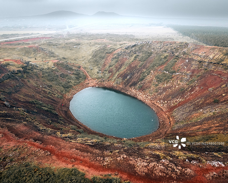 冰岛凯瑞迪火山湖的鸟瞰图，展示了其充满活力的蓝绿色湖水，周围环绕着红色的火山岩和茂密的绿色植被，天空多云图片素材