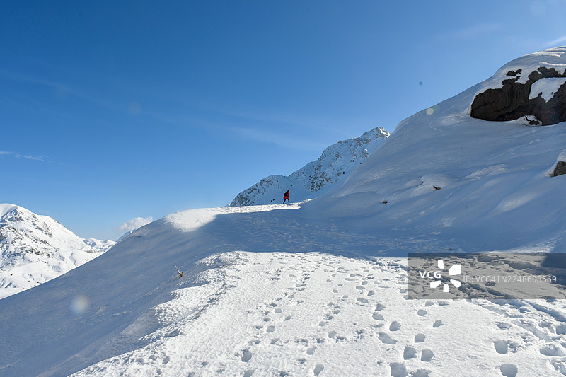 一个穿着红色衣服的人在晴朗的蓝天下，独自走过白雪皑皑的山地景观，身后留下了脚印，土耳其安塔利亚图片素材