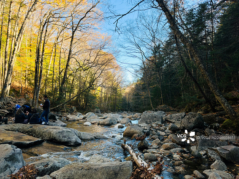 人们在色彩斑斓的森林里，沿着岩石河岸享受着风景如画的秋日时光图片素材