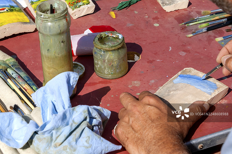 一位艺术家的手在普罗旺斯（法国）的桌子上用画笔在石头上绘画，周围散落着颜料、画笔和眼镜图片素材