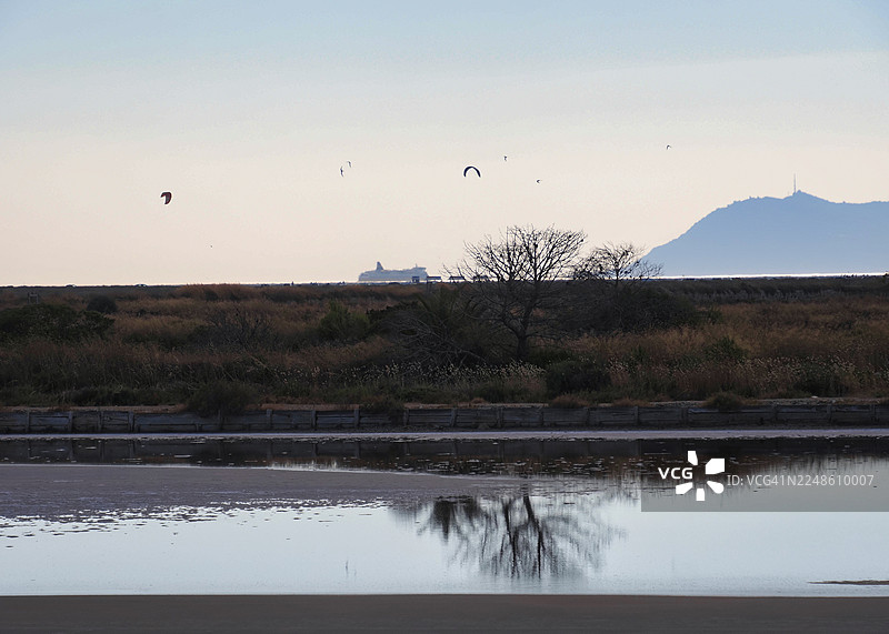 宁静的湿地景观，有倒影、远处的风筝冲浪者、一艘船以及柔和天空下的山，地点：法国拉马德拉格图片素材