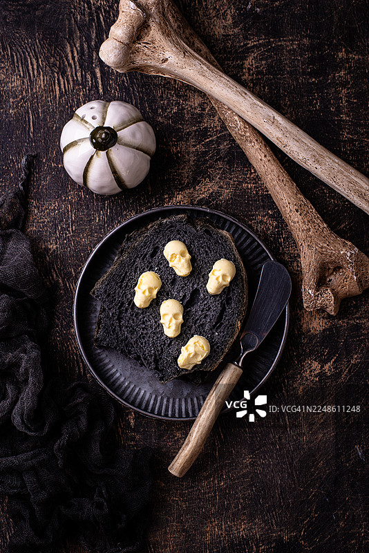 万圣节惊悚吐司，配以骷髅形状的黄油和装饰骨头，摆放在深色桌子上图片素材