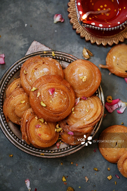高角度拍摄的印度传统甜点，巴卢沙希（Balushahi）或奇罗提（Chirote），盛放在盘子里，配有玫瑰花瓣和开心果，旁边点着一盏迪亚灯（Diya）。图片素材