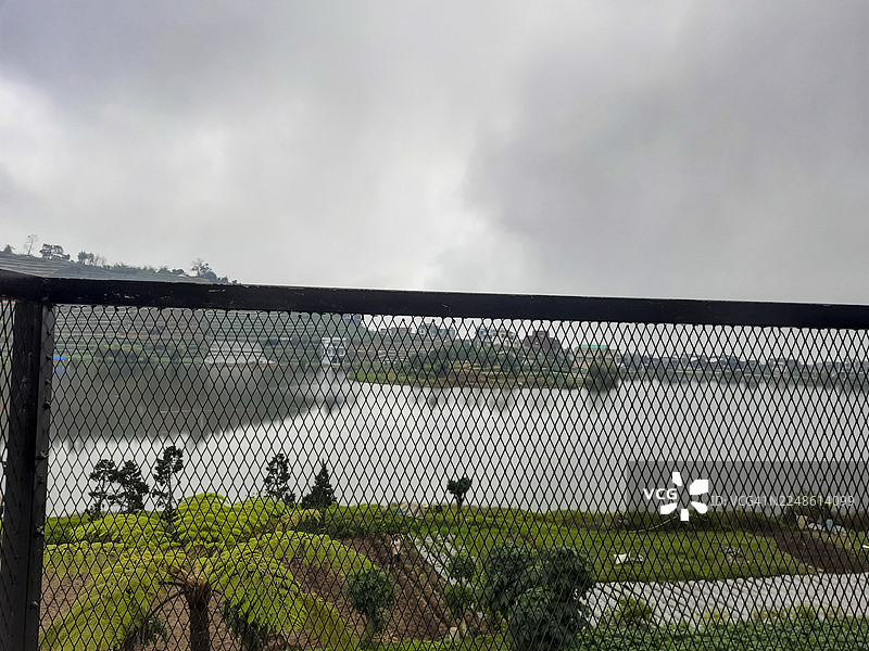 仰视金属网状栅栏，背景是多云的天空，俯瞰着宁静的湖泊和远处郁郁葱葱的山丘。图片素材