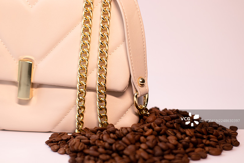 特写镜头：咖啡豆从时尚的米色手提包中溢出，手提包配有金色链条，背景为白色。图片素材