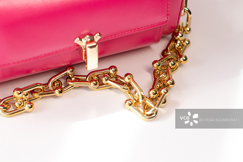 一款奢华时尚的鲜艳粉色钱包特写，配有金色链条和搭扣，背景为纯净的白色。图片素材