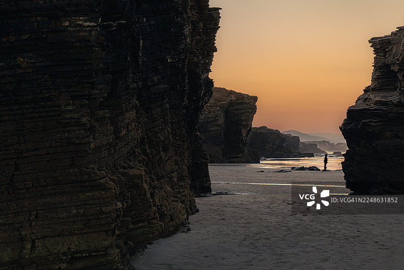 夕阳下，一个人独自站在沙滩上，两旁是高耸的岩石，大海映照着温暖的天空。西班牙，里瓦德奥，大教堂海滩图片素材