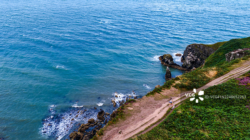 都柏林附近，一位女士带着两只狗沿着俯瞰爱尔兰海的海岸小径在霍斯悬崖步道上行走，景色优美。崎岖的悬崖和绿色的植被，伴随着戏剧性的天空，体现了户外休闲和旅行的生活方式概念。图片素材