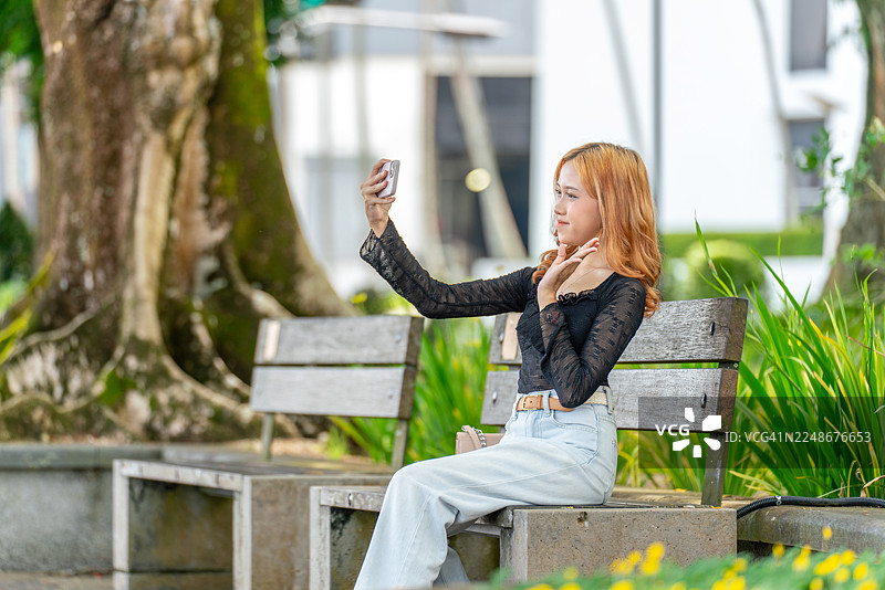 亚洲Z世代女性在古晋滨海公园自拍。年轻的亚洲女性在砂拉越河畔的户外微笑着使用手机。图片素材