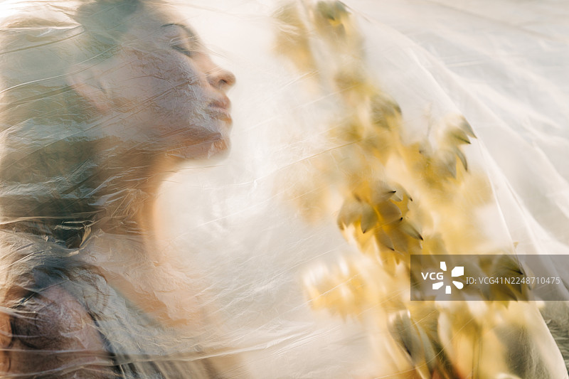 透过一层薄薄的、起皱的塑料薄膜，可以看到一个侧面轮廓的女人和一束黄色的花。这张概念图探讨了环境保护、被压抑的美以及自然与人类影响之间脆弱的界限等主题。图片素材