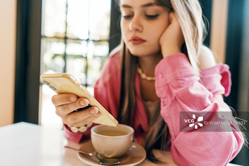 一位穿着粉色衬衫的年轻女子在咖啡馆查看手机，旁边放着一杯咖啡图片素材