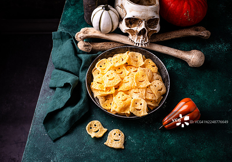 深绿色桌子上的碗里盛着万圣节主题的幽灵形状薯片，旁边装饰着骷髅和南瓜图片素材